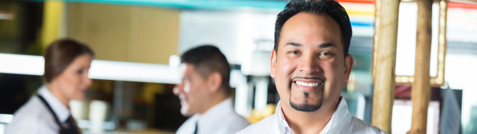 Photo of a man wearing a chef uniform standing in a restaurant. He is smiling and holding a menu.