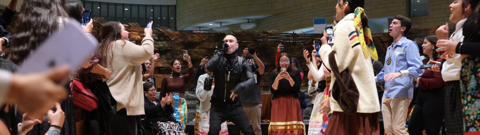 A crowd of Indigenous people wearing traditional attire gather around a singer who is jumping up in the air and holding a mic. He is wearing all black. The people surrounding him are also jumping, smiling, clapping, and being joyful. They are on a light brown floor.
