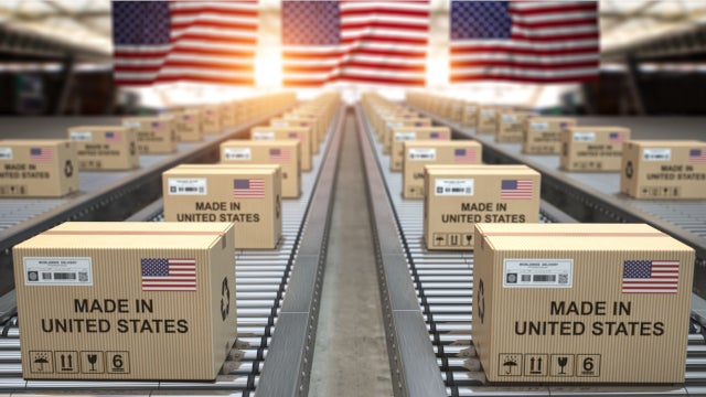 Photo of cardboard boxes moving down a conveyor belt. Each box is stamped with the words "Made in America." There are several American flags hanging in the background.