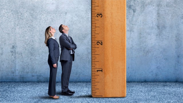A businesswoman and businessman stand at a profile to the camera as they looks up at a large wooden ruler that is standing on its end inside a large room.