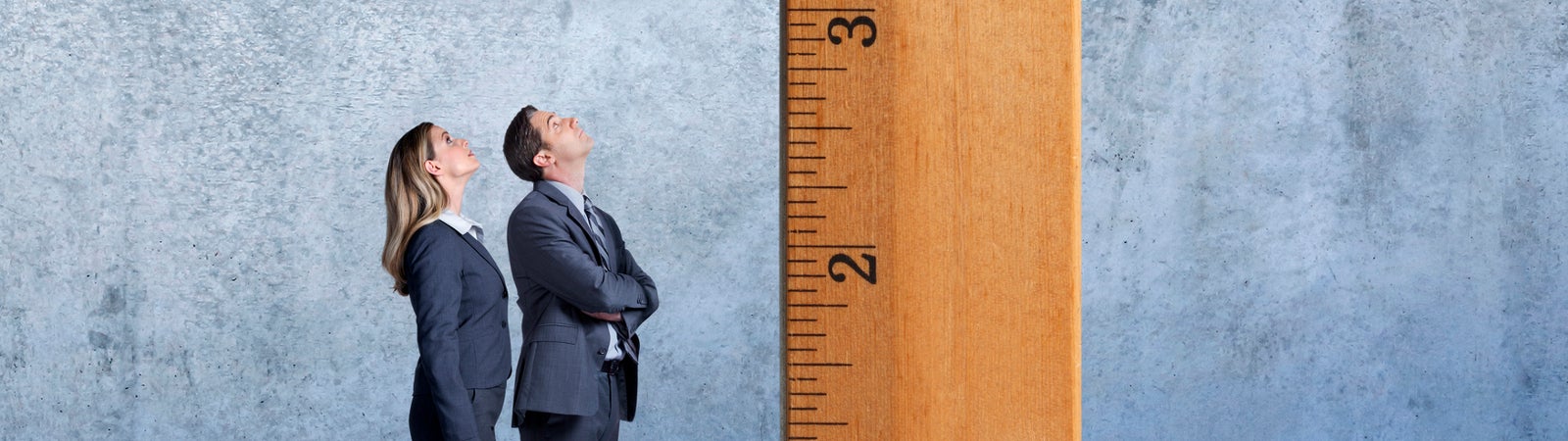 A businesswoman and businessman stand at a profile to the camera as they looks up at a large wooden ruler that is standing on its end inside a large room.